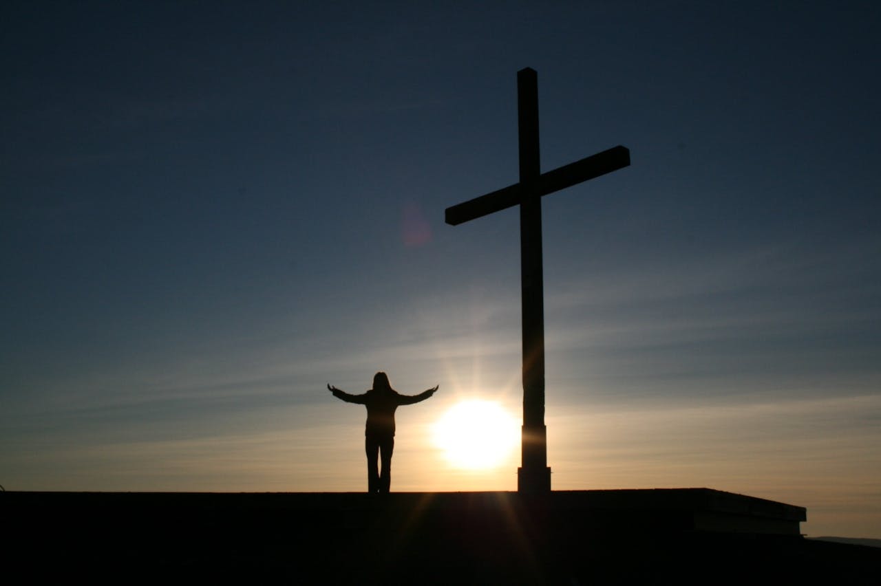 services-01 Silhouette of a person with open arms beside a cross at sunset, symbolizing faith and spirituality.